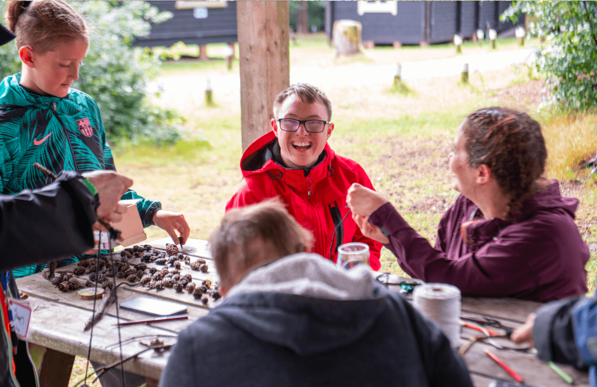 Group of young people sat on a picnic bench making nature arts and crafts in the forest with grass in background