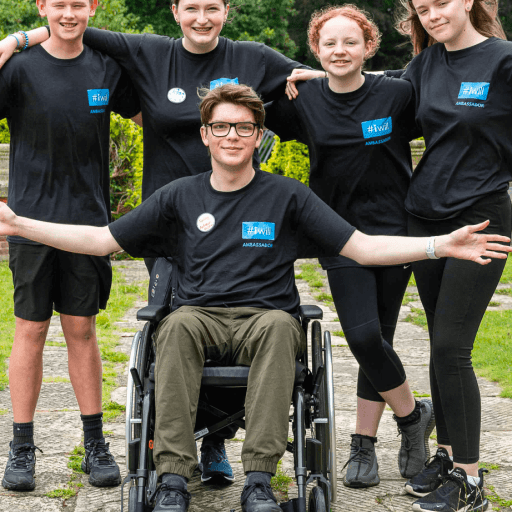 Group of young people with one boy in a wheelchair smiling to camera on a pathway in the forest some with open arms and others with arms around shoulders