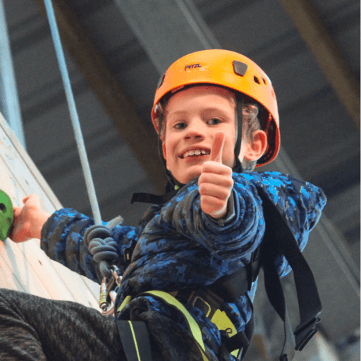 Young boy at the top of a climbing wall smiling to camera with a harness attached to a rope. Boy is holding thumbs up