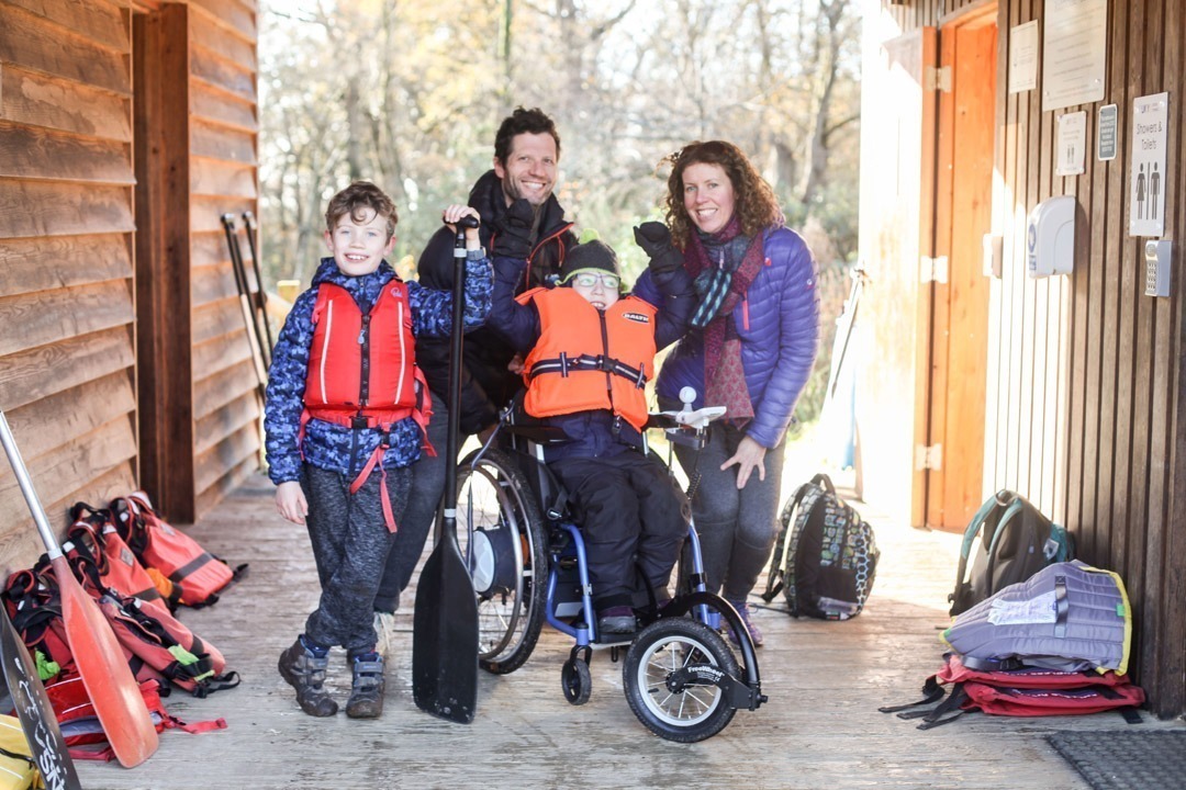 Family of four in a wooden structure with two children wearing life jackets, one of the children is sat in a wheelchair with paddle raised