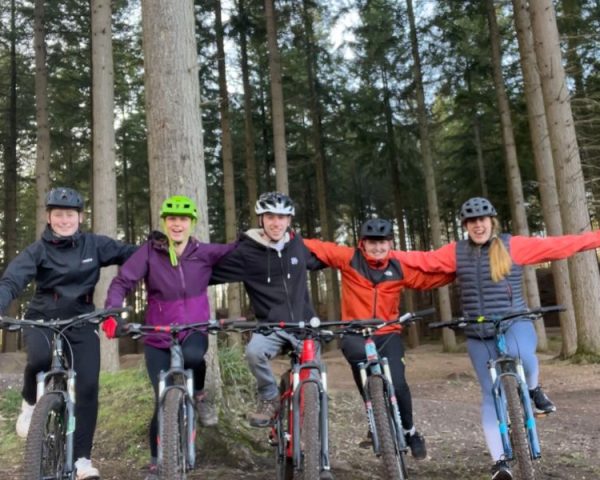 5 adults on bikes with arms on shoulders smiling to camera all wearing helmets