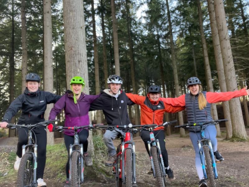 5 adults on bikes with arms on shoulders smiling to camera all wearing helmets