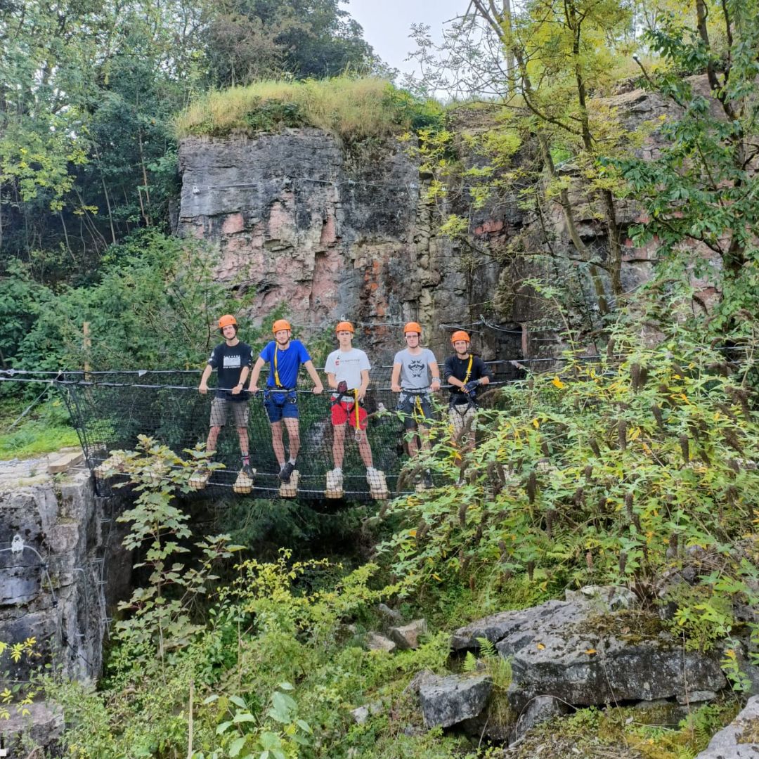 5 male adults stood on netted bridge i the forest traversing