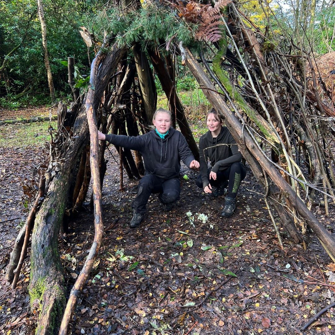 Two adults smiling in a natural shelter made from branches, twigs and leaves