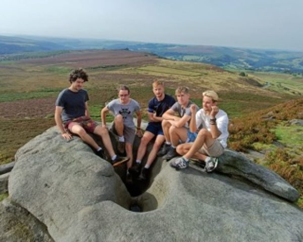5 male adults sat on a big rock smiling to camera in front of scenic view of countryside
