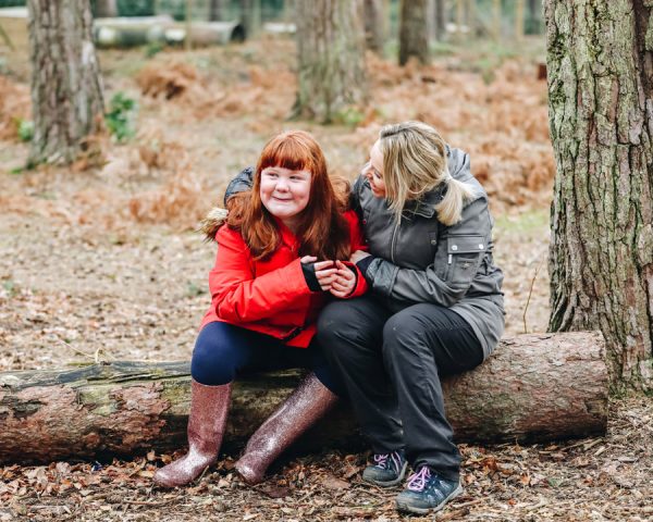Young girl and female adult hugging and smiling sat on a wooden branch in the middle of a forest in the winter