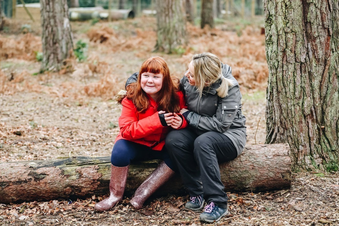 Young girl and female adult hugging and smiling sat on a wooden branch in the middle of a forest in the winter
