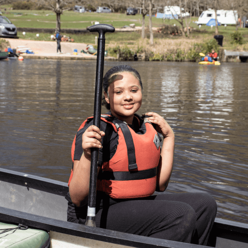 Young person sat in a canoe smiling to camera wearing a life jacket and holding an oar