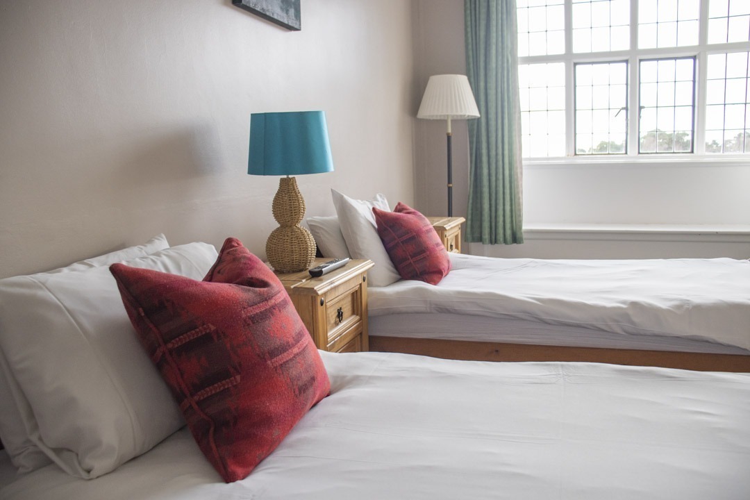 Inside the Avon Tyrell manor house - bedroom with two single beds and window - both beds have a red pillow that is decorative and two little lamps and small side table