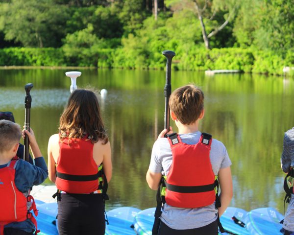 Four young people lined up looking away from camera towards a still lake with trees in the background. All young people are wearing life jackets and holding paddles