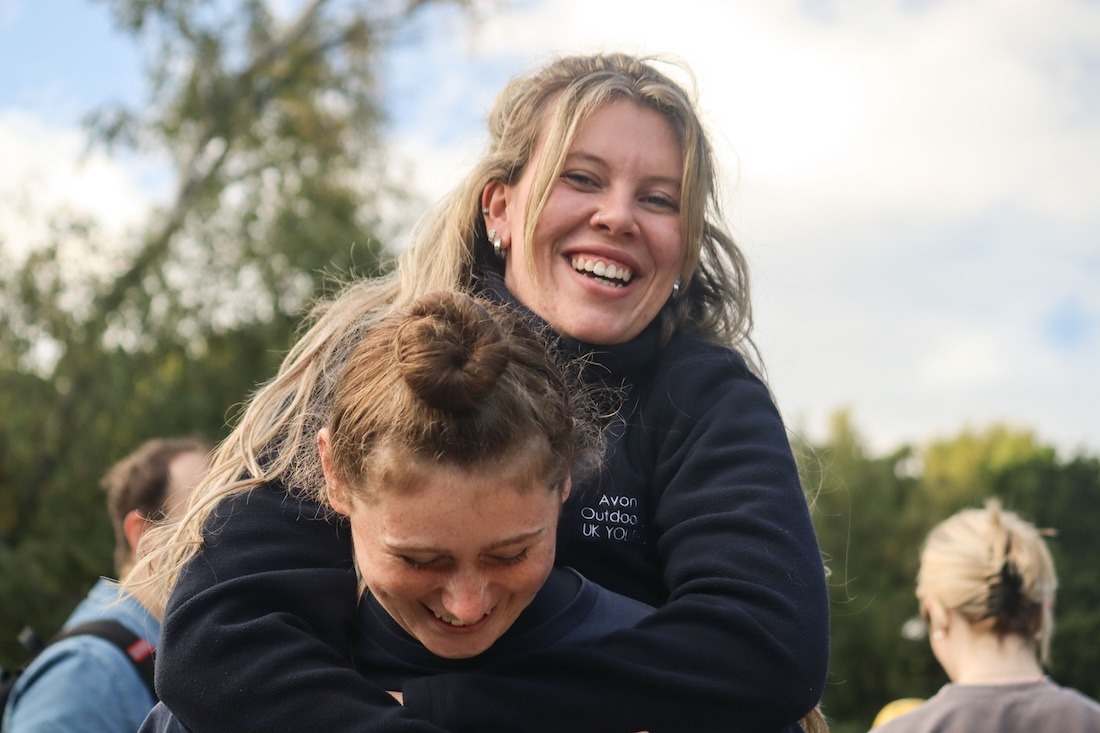 Two instructors smiling to camera with one giving the other piggy back