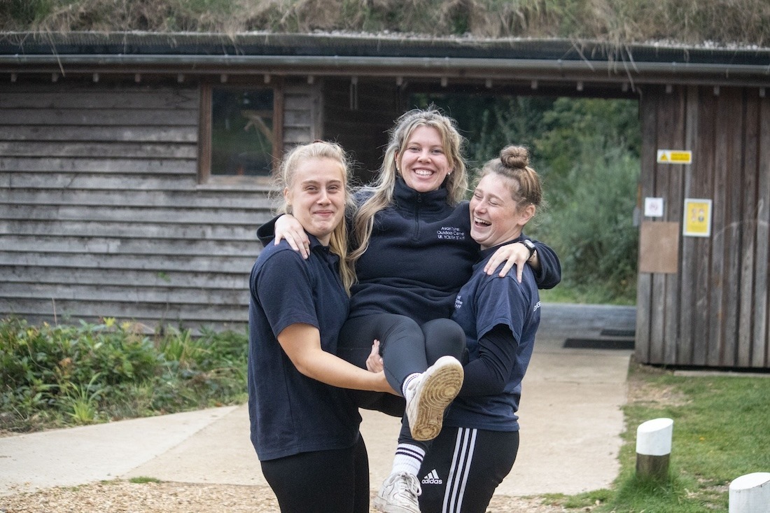 Three adults one being lifted by the two others in front of wooden building in the forest