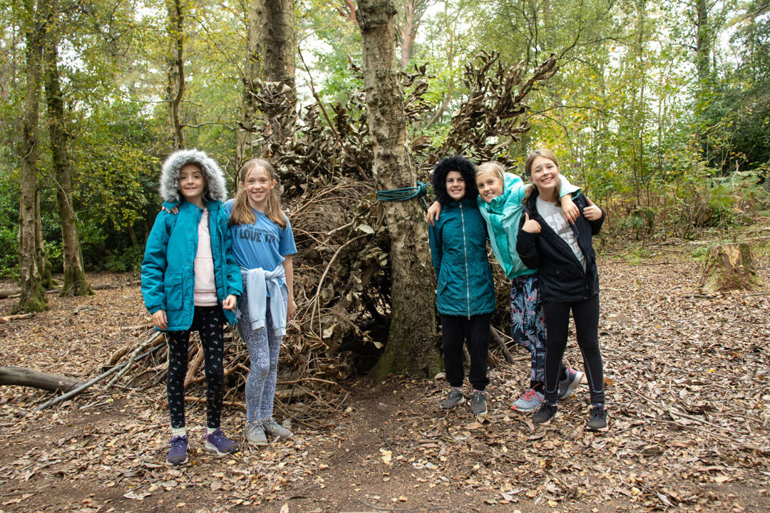 young people stood in forest building shelter out of things, branches and leaves smiling to camera