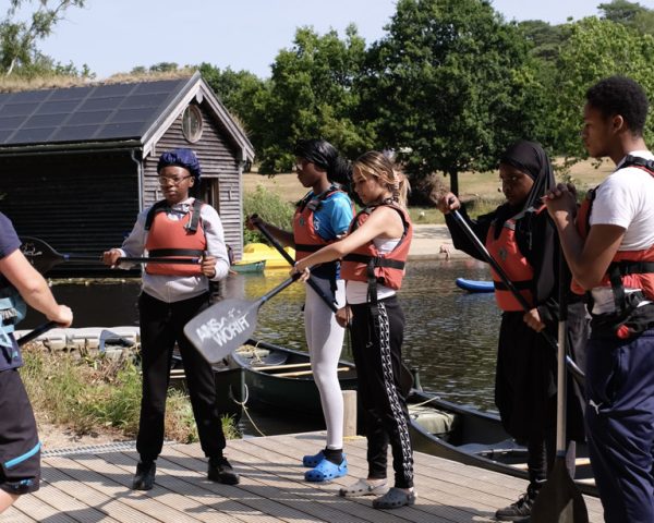 Group of young people stood on wooden structure infront of lake preparing for canoeing session learning how to use oars - all wearing life jackets