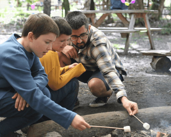 Two boys and man roasting marshmallows over a fire in the forest