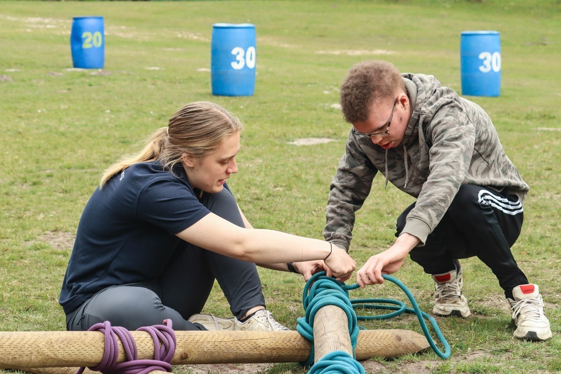 Young person and outdoor instructor at Avon Tyrrell making a catapult out of wooden pieces and rope with blue barrels in the backfground