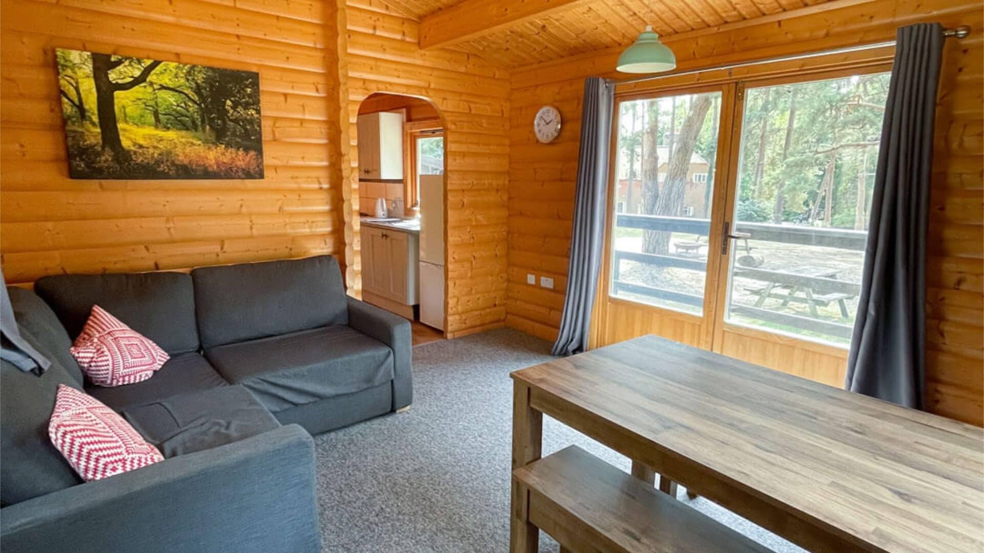 Inside lounge area of an Avon Tyrrell 6 berth lodge in the new forest. There is a grey corner sofa with red pillows, two windows, dining table, wooden benches and TV on the wall. The walls are wooden