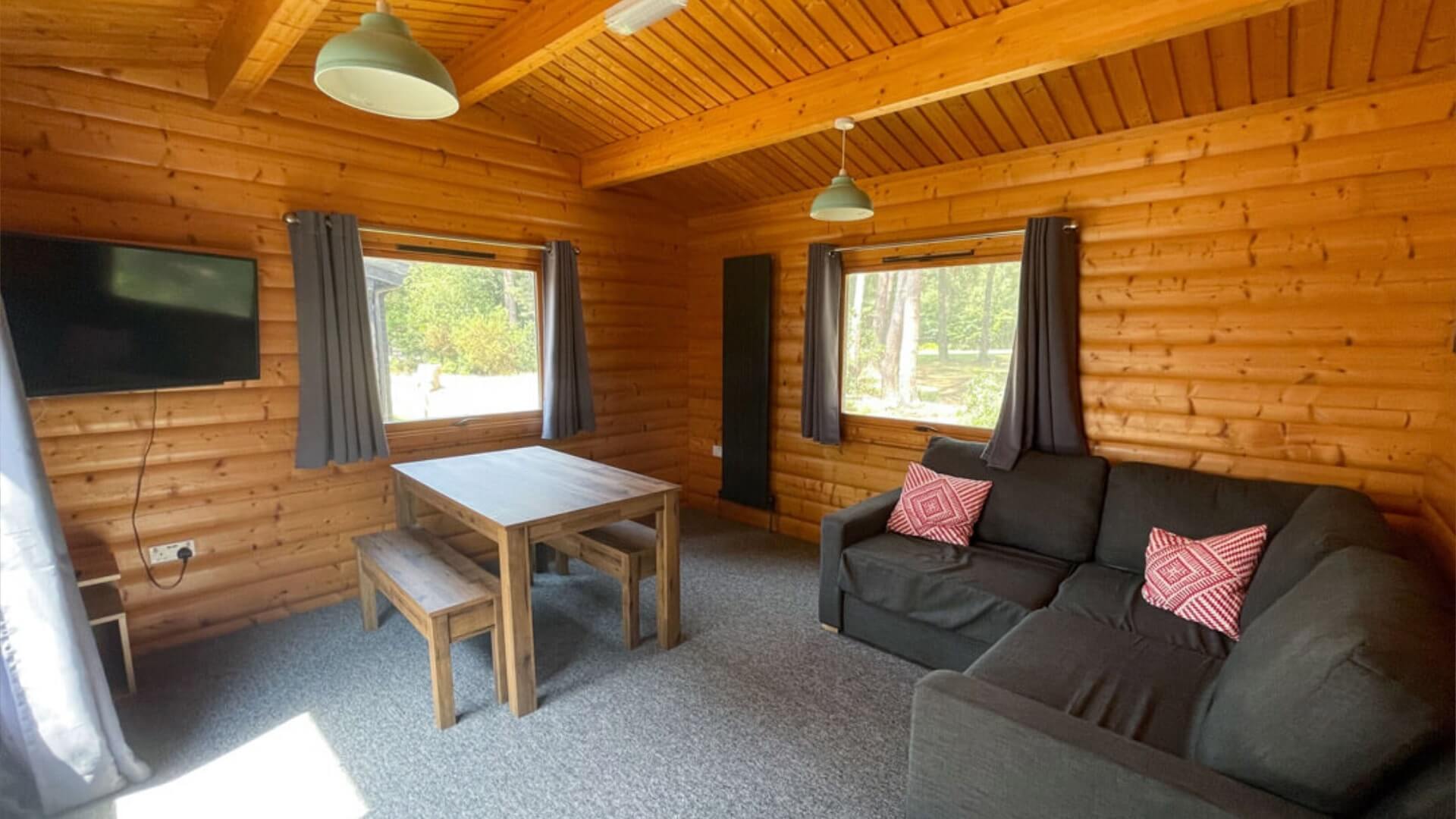 Inside lounge area of an Avon Tyrrell 6 berth lodge in the new forest. There is a grey corner sofa with red pillows, two windows, dining table, wooden benches and TV on the wall. The walls are wooden