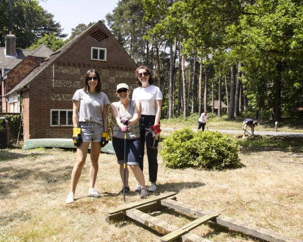 Three adult ladies smiling towards camera with stone building in the background. In a forest with grass on the floor, the people are wearing gardening gloves and gardening forest area