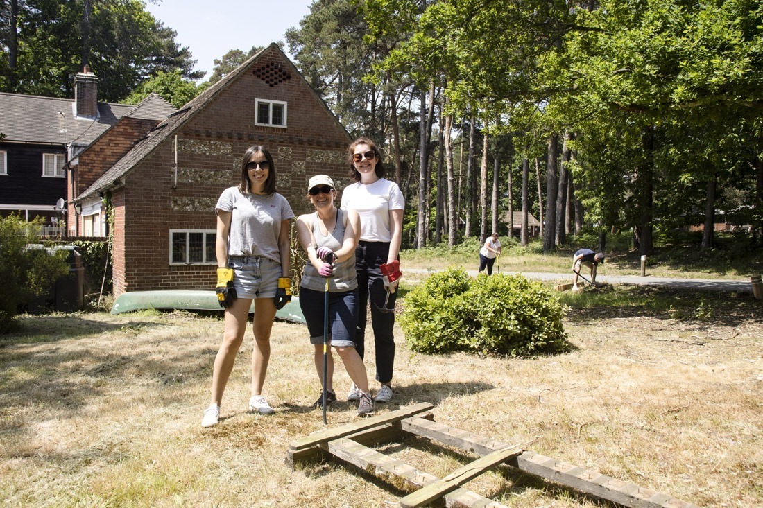 Three adult ladies smiling towards camera with stone building in the background. In a forest with grass on the floor, the people are wearing gardening gloves and gardening forest area