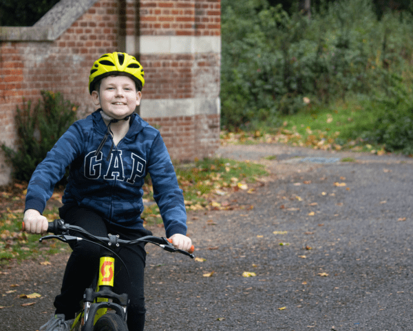 Young person wearing gap hoodie and yellow helmet on a yellow bike smiling to camera with a stone fence