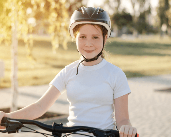 Young girl wearing helmet stood over bike with hands on handles on a pavement with trees and grass in background