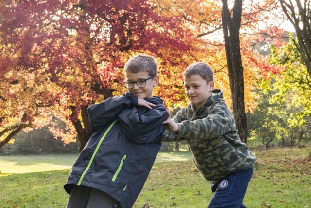 Two young boys stood in forest with autumnal trees in background - one boy is leaning back to the other boy