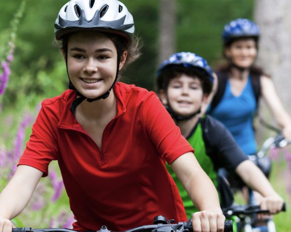 Young women in focus at front of image cycling on a bike through flower field wearing a helmet - younger boy and adult cycling wearing helmets behind