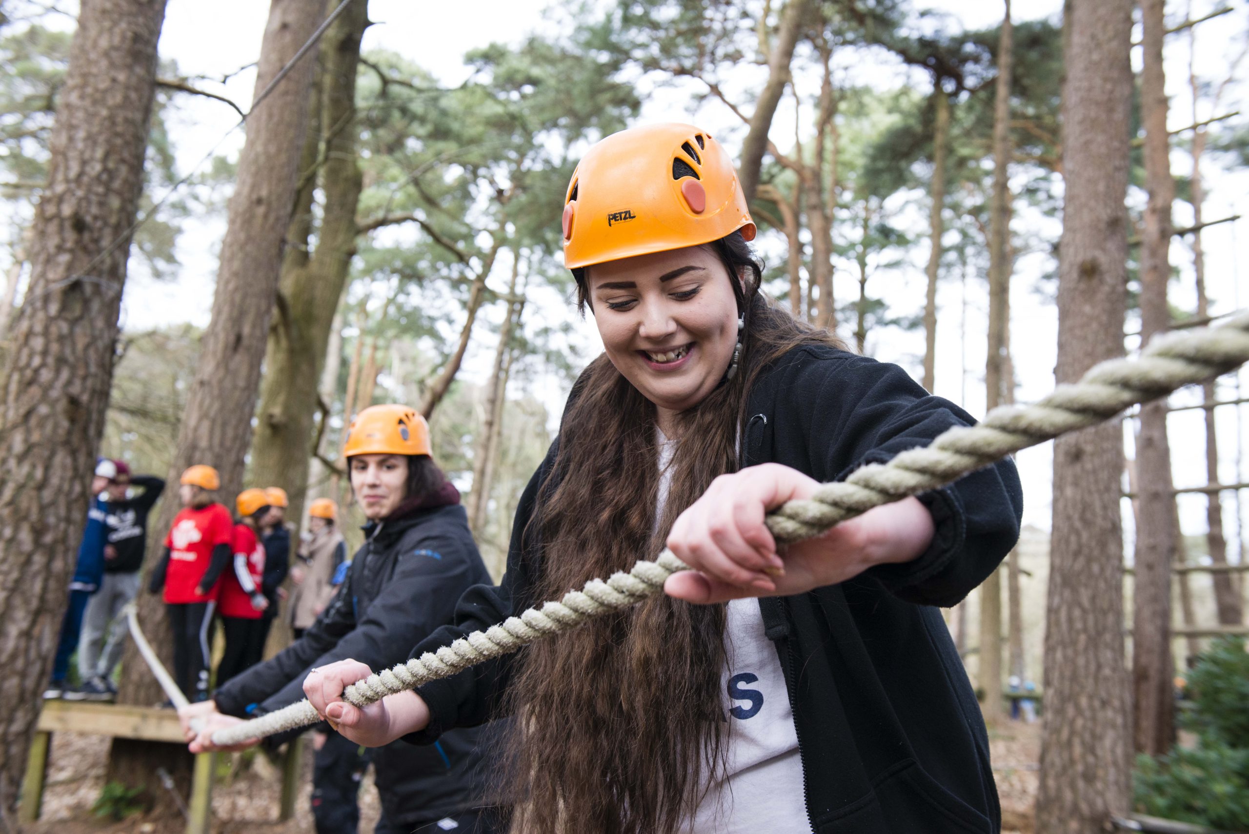 Young adult wearing adult smiling looking down walking with rope guide in her hand. person behind her is doing the same both people are wearing helmets