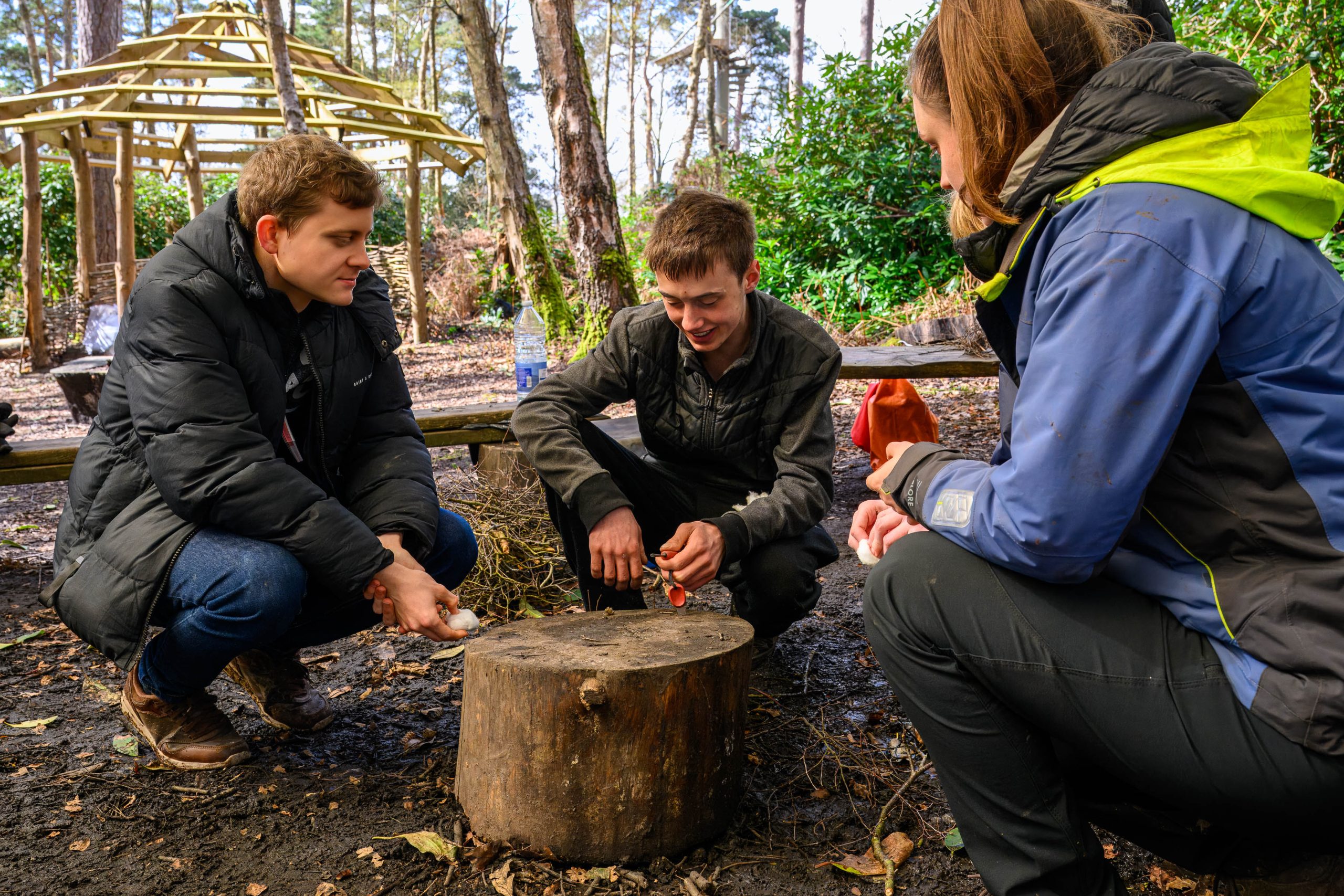 Two young adult males with feamale Avon Tyrrell instrutor knelt next to wooden stump in forest trying to light with flint and steel