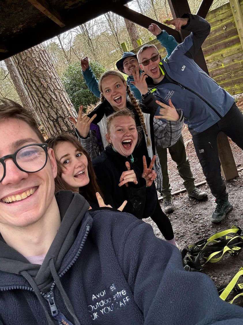 Apprentices smiling to camera under a shelter with climbing harnesses in the New Forest at Avon Tyrrell Outdoor Centre