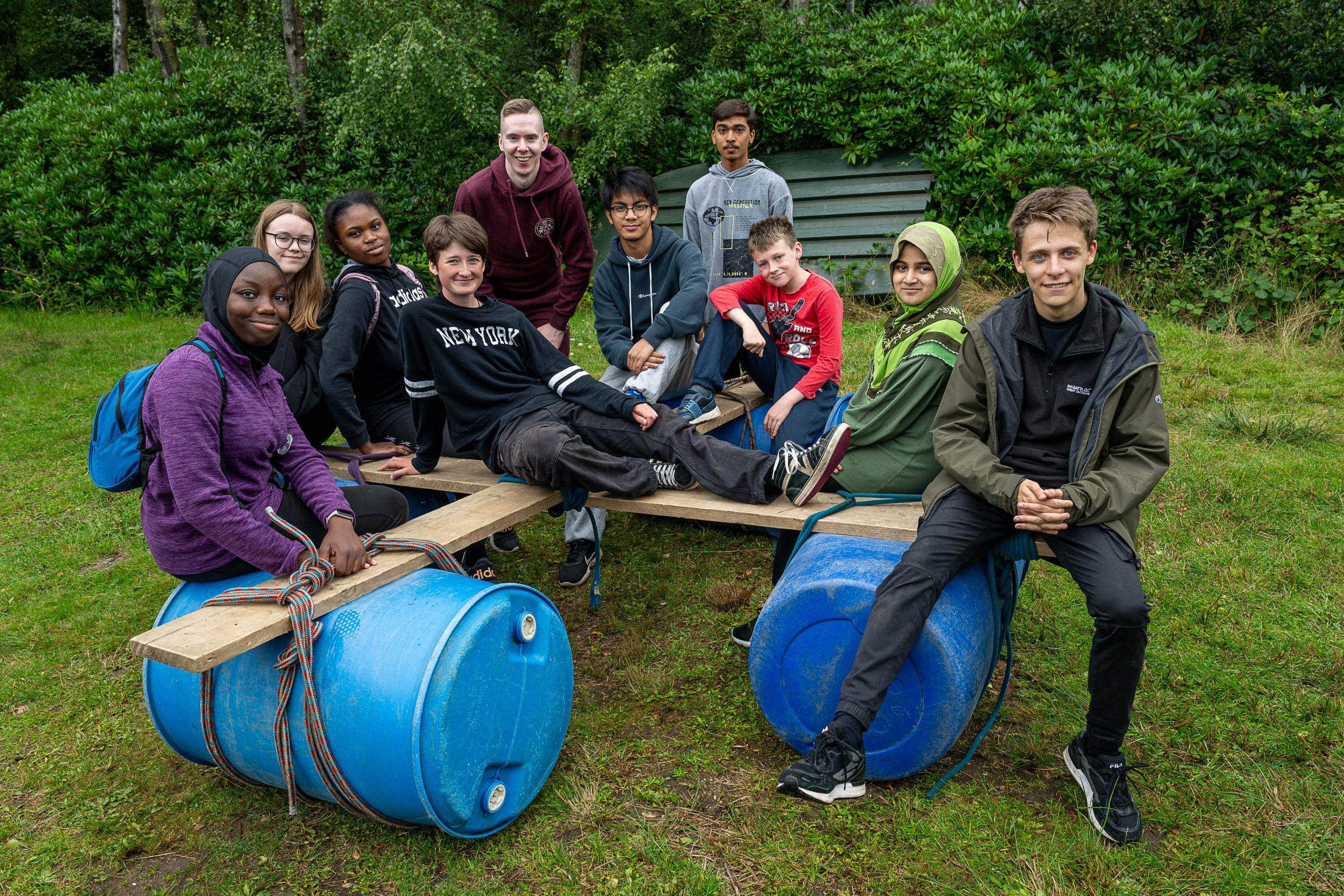 Group of young people sat on a hand made wooden raft with a mix of male and female. Raft is made from blue barrels, wood and ropes