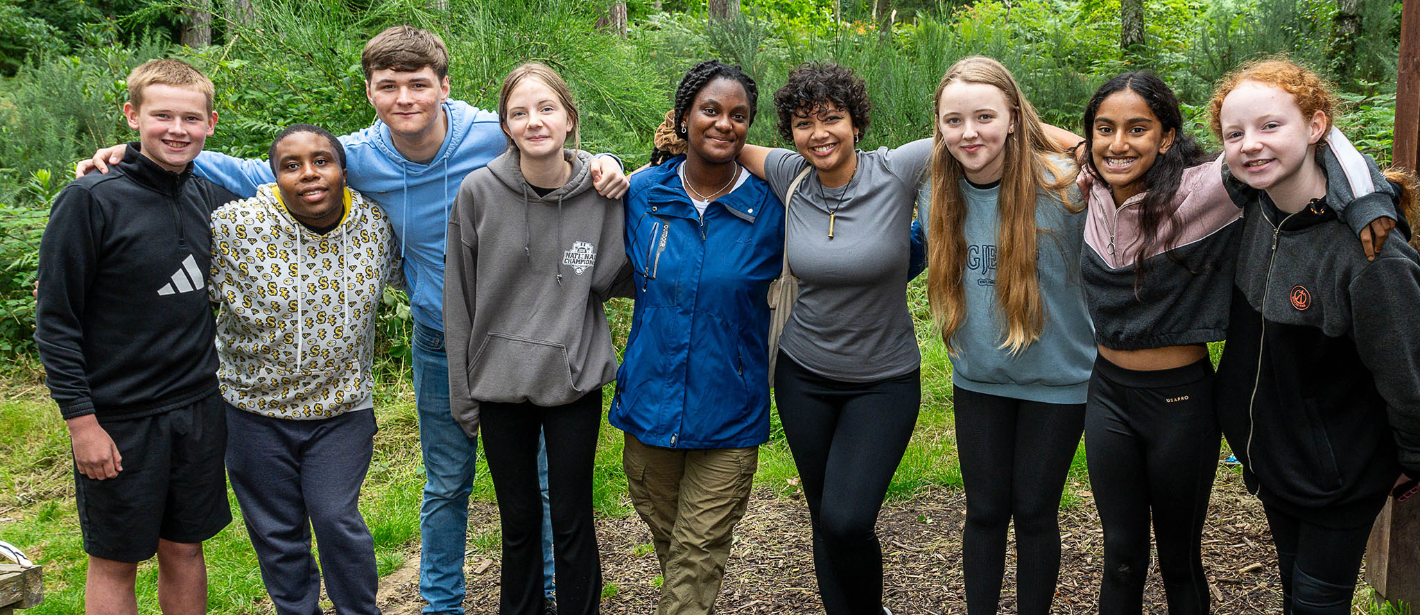 Group of young people stood in the forest with arms around each others shoulders smiling to camera